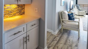 A modern kitchen with white cabinets and a brick backsplash, leading to a living area with a white armchair and a view of the outdoors. - Ocean Creek Kk-14 2 Bedroom Condo by RedAwning (Myrtle Beach)