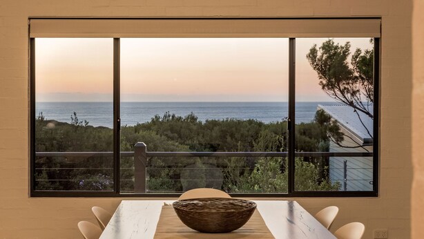 A dining area with a view of the ocean, a large window, and a wicker bowl on the table.