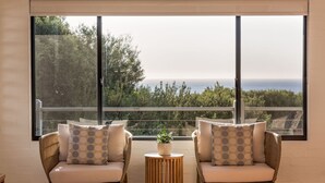 A room with two wicker chairs, a small round wooden table with a potted plant, and a large window offering a view of the sea and greenery.