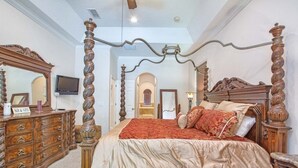 Una cama con dosel con colcha roja, un aparador de madera con varios cajones, un televisor en la pared y un espejo.