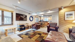 A modern living room with a flat-screen TV, leather sofas, a coffee table, and a kitchen area in the background.