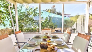 Mesa de comedor preparada para cuatro personas, con cubierta de vidrio, rodeada de sillas blancas con cojines marrones, y vistas a zonas verdes y un edificio a través de grandes ventanales.