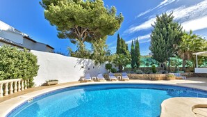 Una piscina con sillones de descanso, rodeada de árboles y una pared blanca.