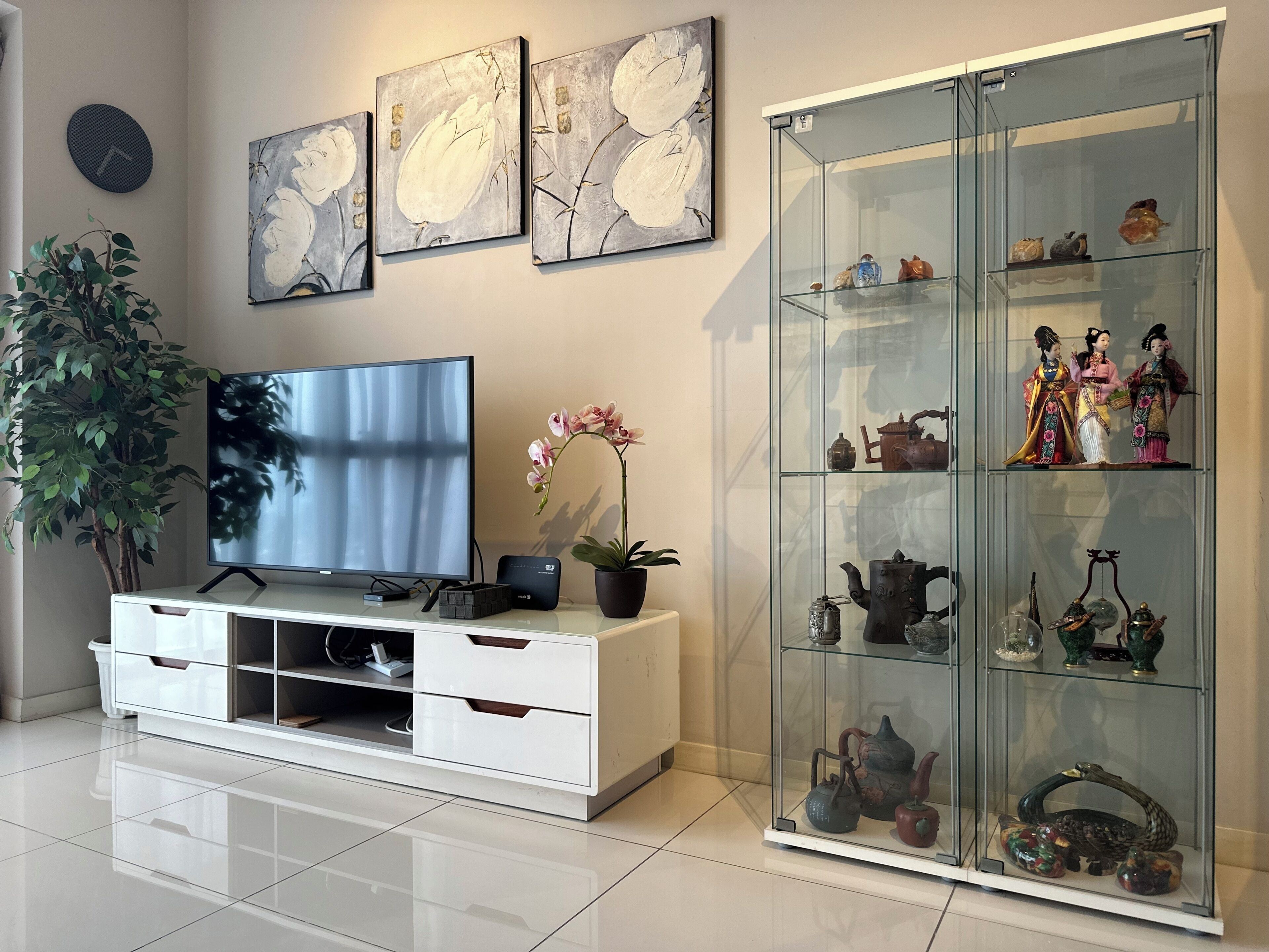 Una sala moderna con un televisor de pantalla plana, un aparador de vidrio con adornos y una planta en maceta.