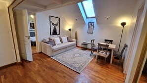 A modern living room with a sofa, a dining table, and chairs. There is a large window with a skylight, a framed picture on the wall, and a lamp. - TyzenXL Luxury Vilnius Apartment (Vilnius)