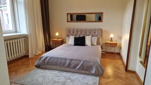 A bedroom with a tufted headboard, a large bed with a patterned bedspread, two bedside tables with lamps, a mirror above the bed, and a window with curtains. - TyzenXL Luxury Vilnius Apartment (Vilnius)