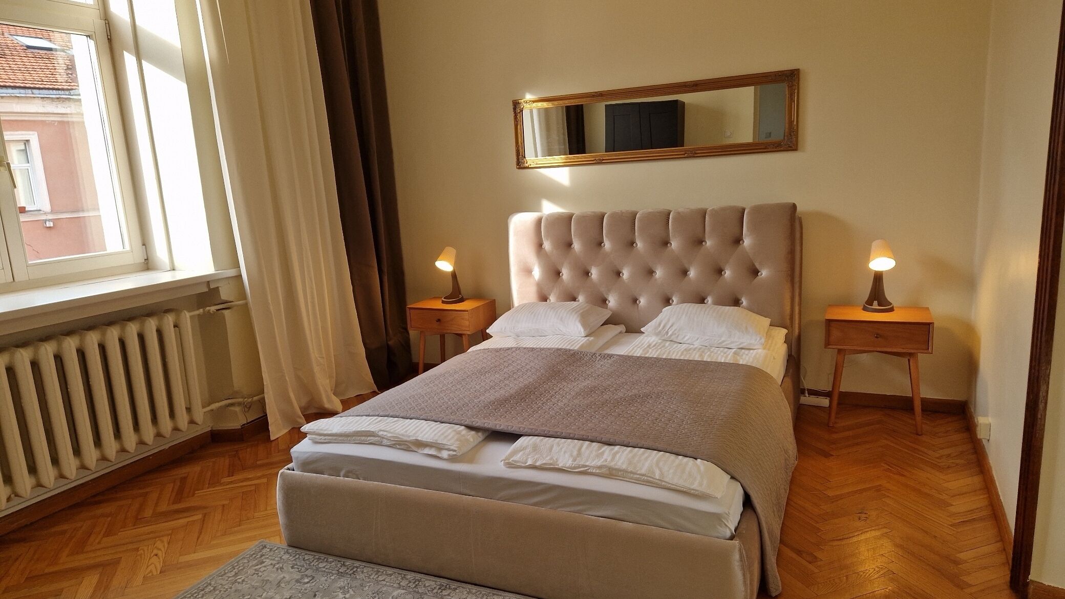 Une chambre à coucher bien rangée, avec un grand lit, deux tables de chevet avec des lampes, un miroir au-dessus du lit et un radiateur sur le côté gauche.