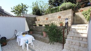 A stone-floored patio with a white table and chairs, potted plants, and a stone wall with steps. - Apartment Irena (Pula)