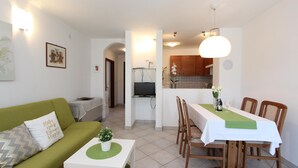 A compact living space with a green sofa, a dining table with chairs, a small white table, and a kitchen area with wooden cabinets and a TV. - Apartment Irena (Pula)