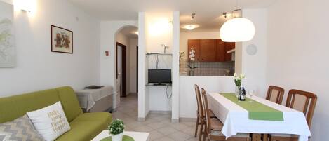 A compact living space with a green sofa, a dining table with chairs, a small white table, and a kitchen area with wooden cabinets and a TV.