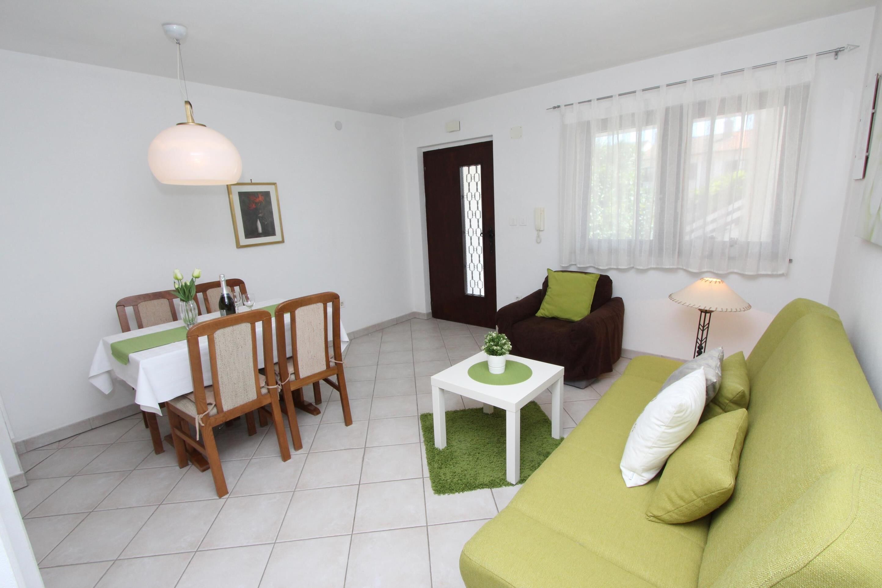 Una sala de estar con un sofá verde, una mesa de comedor con sillas, una mesita blanca con una planta y una lámpara.