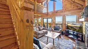 A spacious wooden interior with a balcony, a ceiling fan, and a large TV screen displaying a scenic view. - Endless VIEWS! 3 decks - cozy - Private-with Game Room, Hot Tub, Arcade (Pigeon Forge)