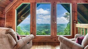 A wooden cabin with a large window offering a view of green hills and mountains. - Endless VIEWS! 3 decks - cozy - Private-with Game Room, Hot Tub, Arcade (Pigeon Forge)