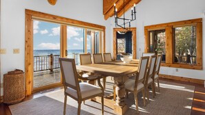 A wooden dining table with six chairs, large windows with ocean views, and a wooden ceiling.