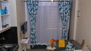 A compact hotel room with a kitchenette, a sofa, a bed, and a small table with flowers.