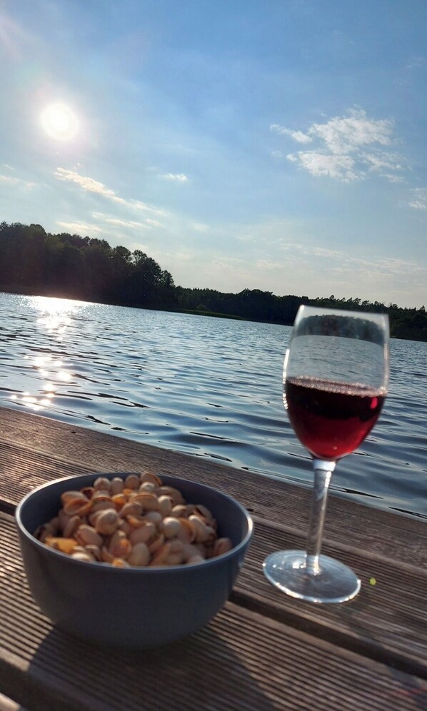 A glass of red wine and a bowl of nuts on a wooden surface by a lake.