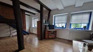 Una habitación amplia con suelo de madera, un mueble empotrado para televisión y dos ventanas con persianas.