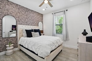 A bedroom with a bed, two mirrors, a ceiling fan, a window with curtains, and a bedside table with a vase.