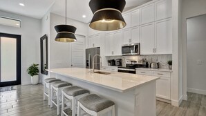 A modern kitchen with white cabinetry, a large island with bar stools, stainless steel appliances, and pendant lighting. - Golf Course Views, Private Pool & Pet-Friendly (Bonita Springs)