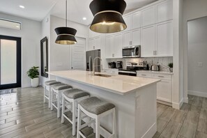 A modern kitchen with white cabinetry, a large island with bar stools, stainless steel appliances, and pendant lighting. - Golf Course Views, Private Pool & Pet-Friendly (Bonita Springs)