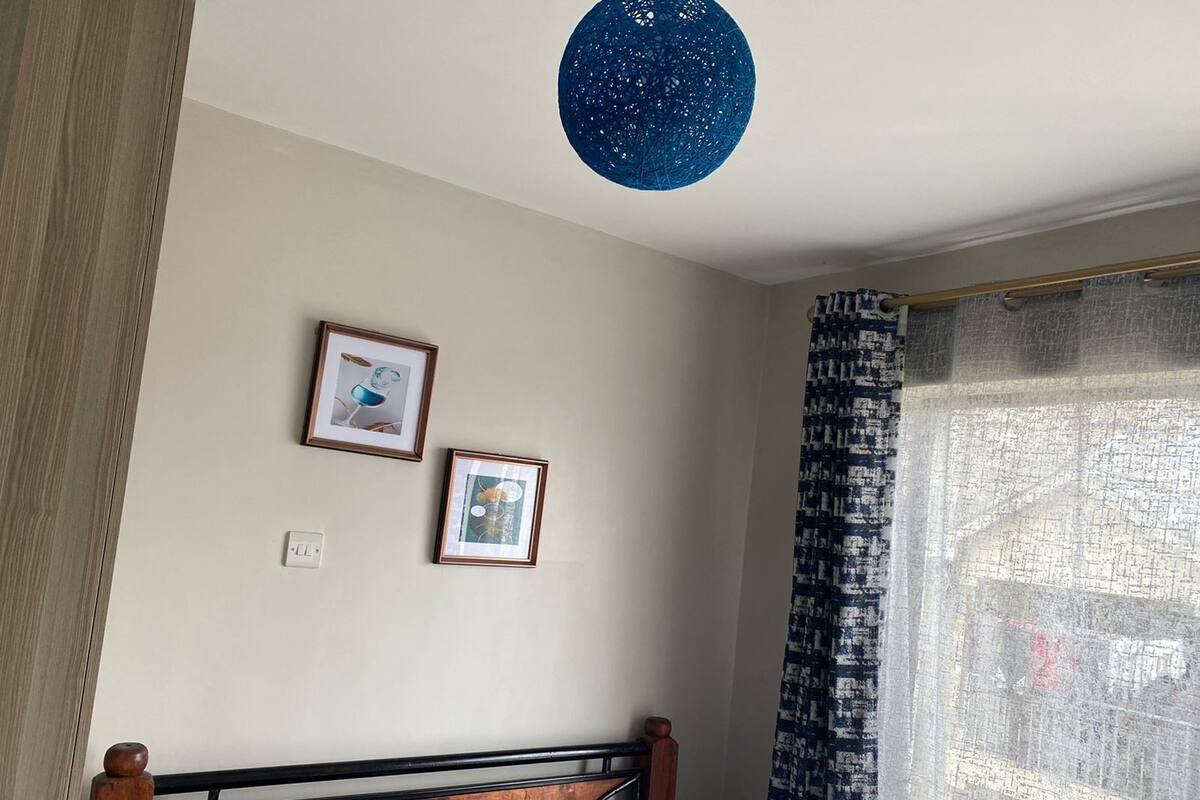 A bedroom with a bed, two framed pictures on the wall, a blue hanging light, and a window with patterned curtains.