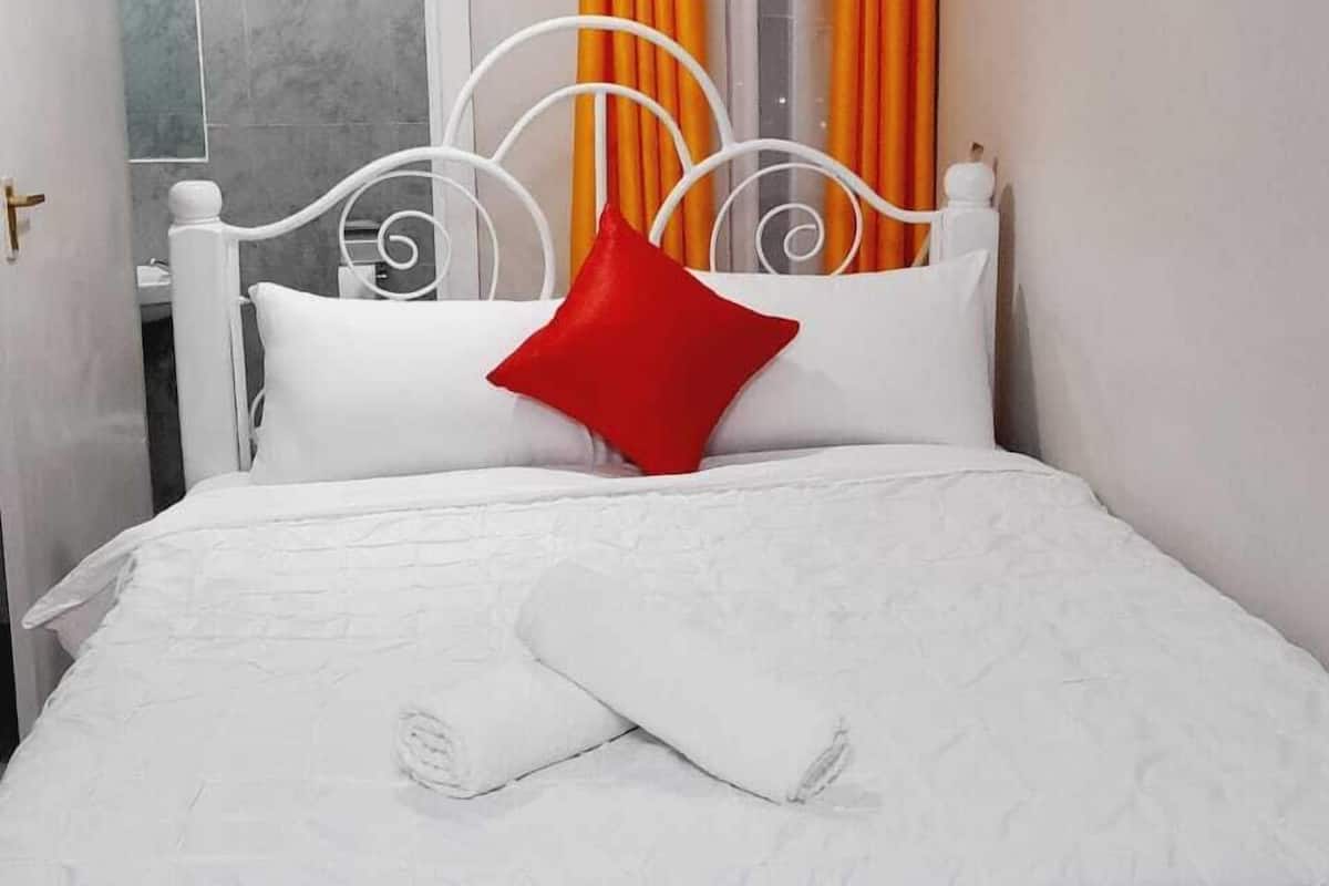 A neatly made bed with white bedding and a red pillow, a white metal headboard, and orange curtains.