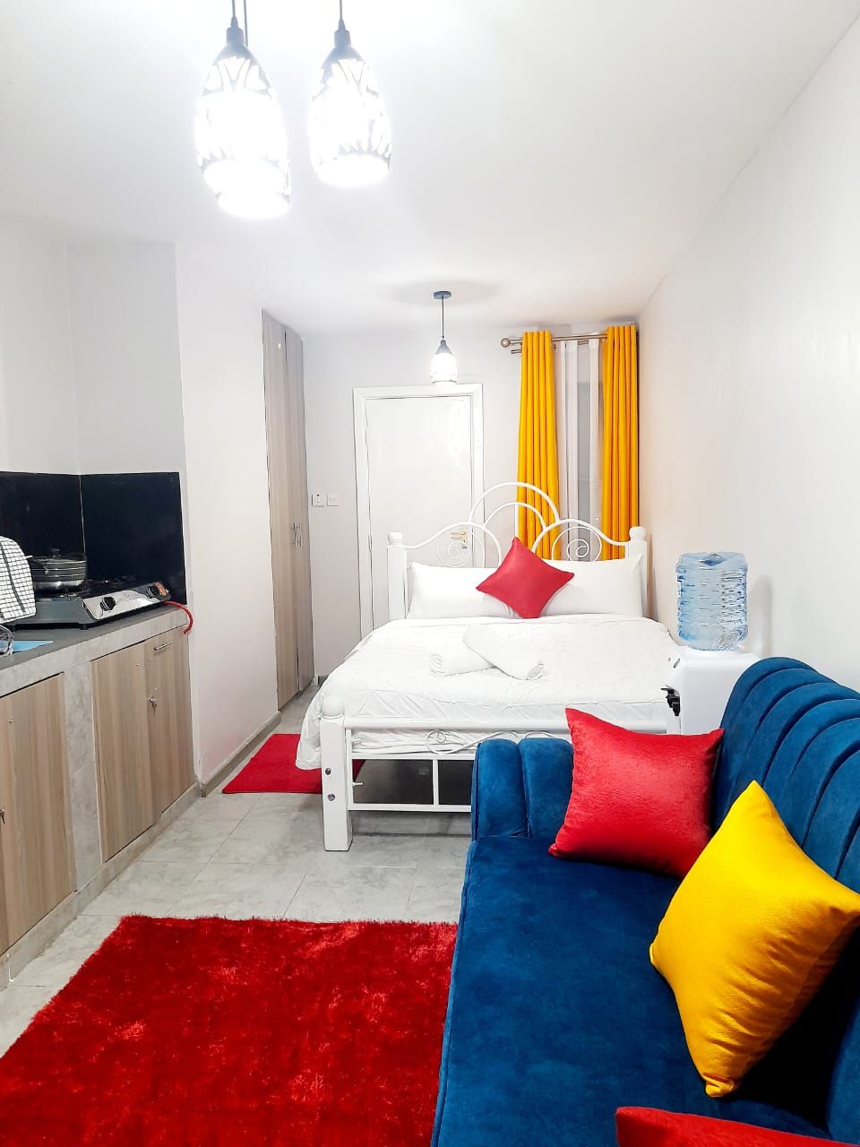 Une chambre à coucher avec un lit, un canapé bleu, un tapis rouge et un distributeur d’eau.