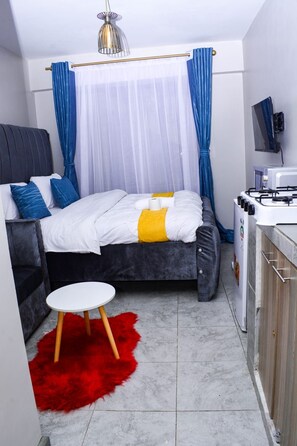 A compact hotel room with a bed, a small round table, a red furry rug, a kitchenette, and a television. - Lux Suites Convertible Studios Embakasi (Nairobi)