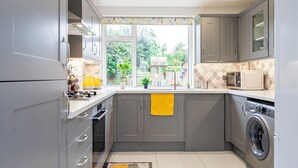 A compact kitchen with grey cabinets, a washing machine, and a window with a view of a garden. - Stunning Place near Windsor & Heathrow, 3BR House (Burnham)