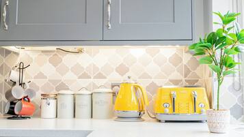 Une cuisine avec des meubles gris, un plan de travail blanc et un crédence à motifs. On y trouve deux appareils de couleur jaune, une bouilloire et un grille-pain.