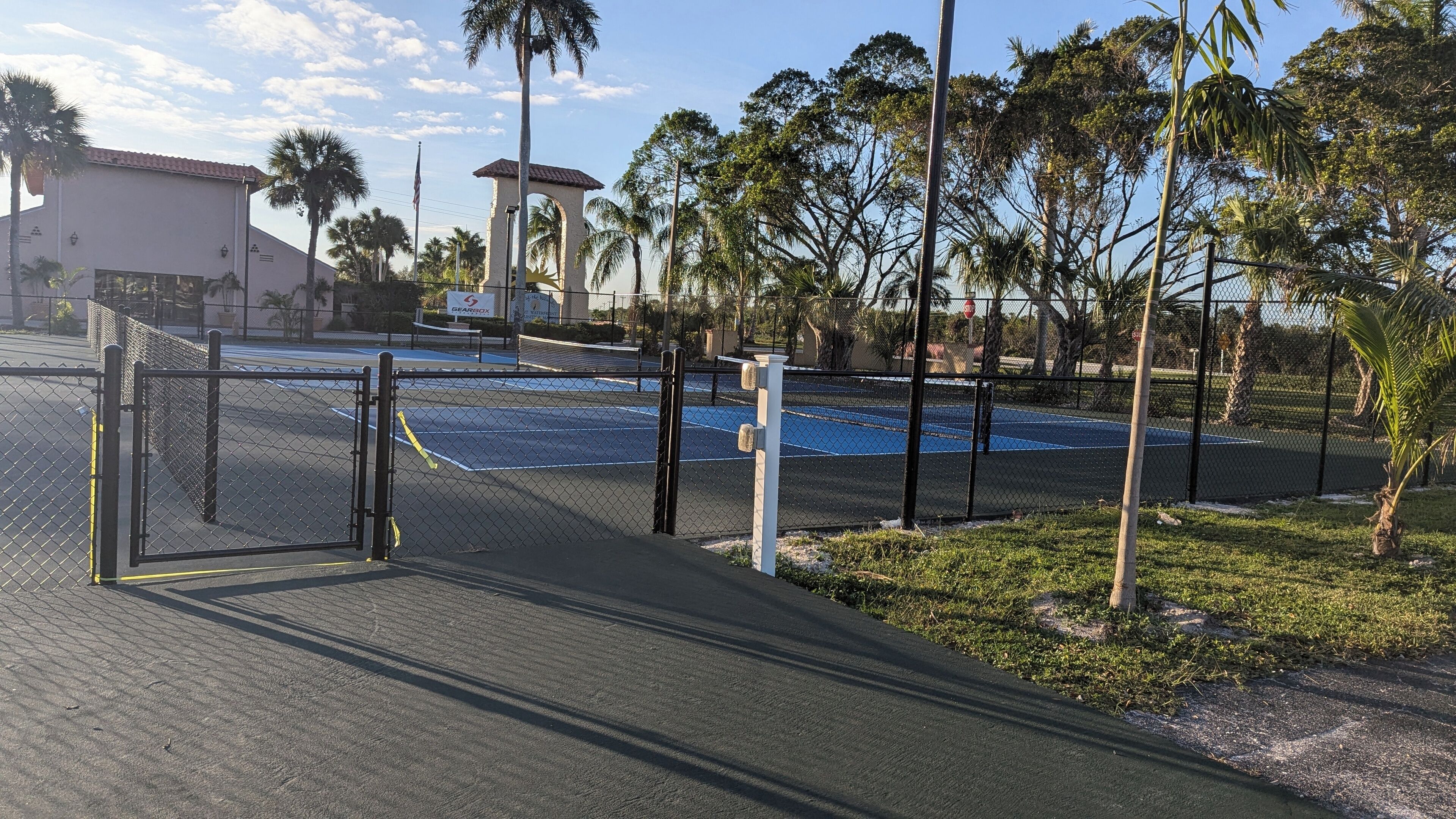 Ett inhägnat parkeringsområde med en blå tennisbana, palmer och en byggnad med ett rött tak i bakgrunden.