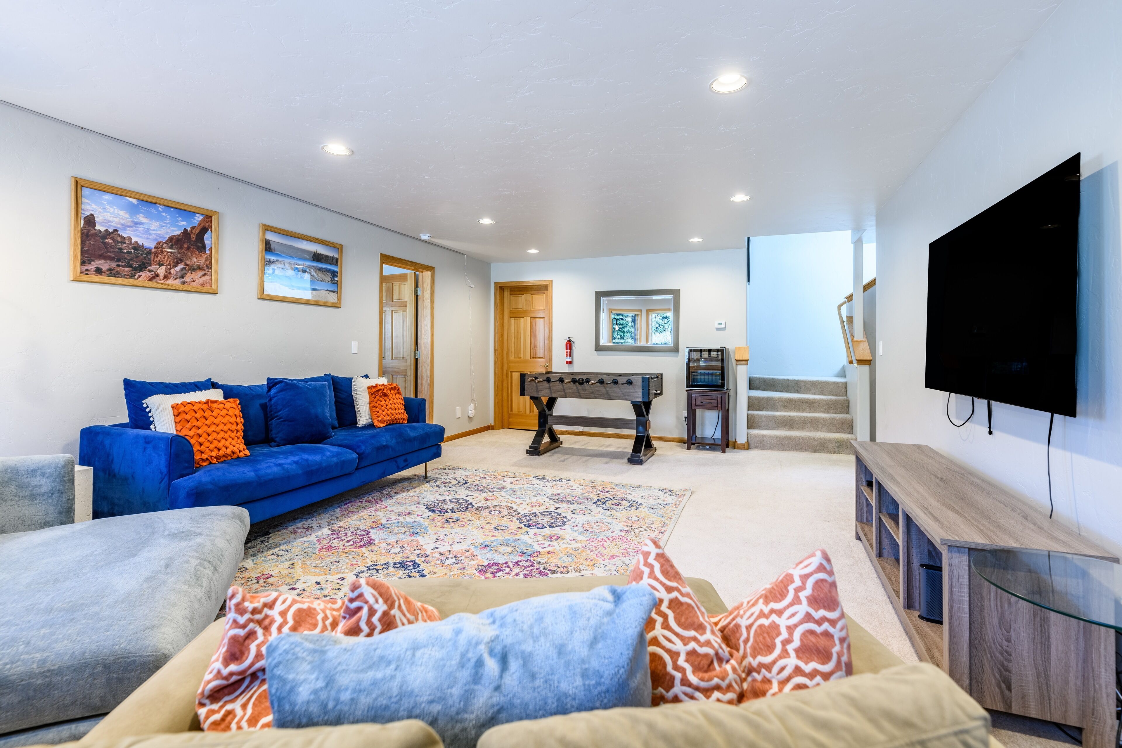 A spacious living room with a blue sofa, a beige sofa, and a coffee table. There is a foosball table, a television, and a staircase leading to another level.