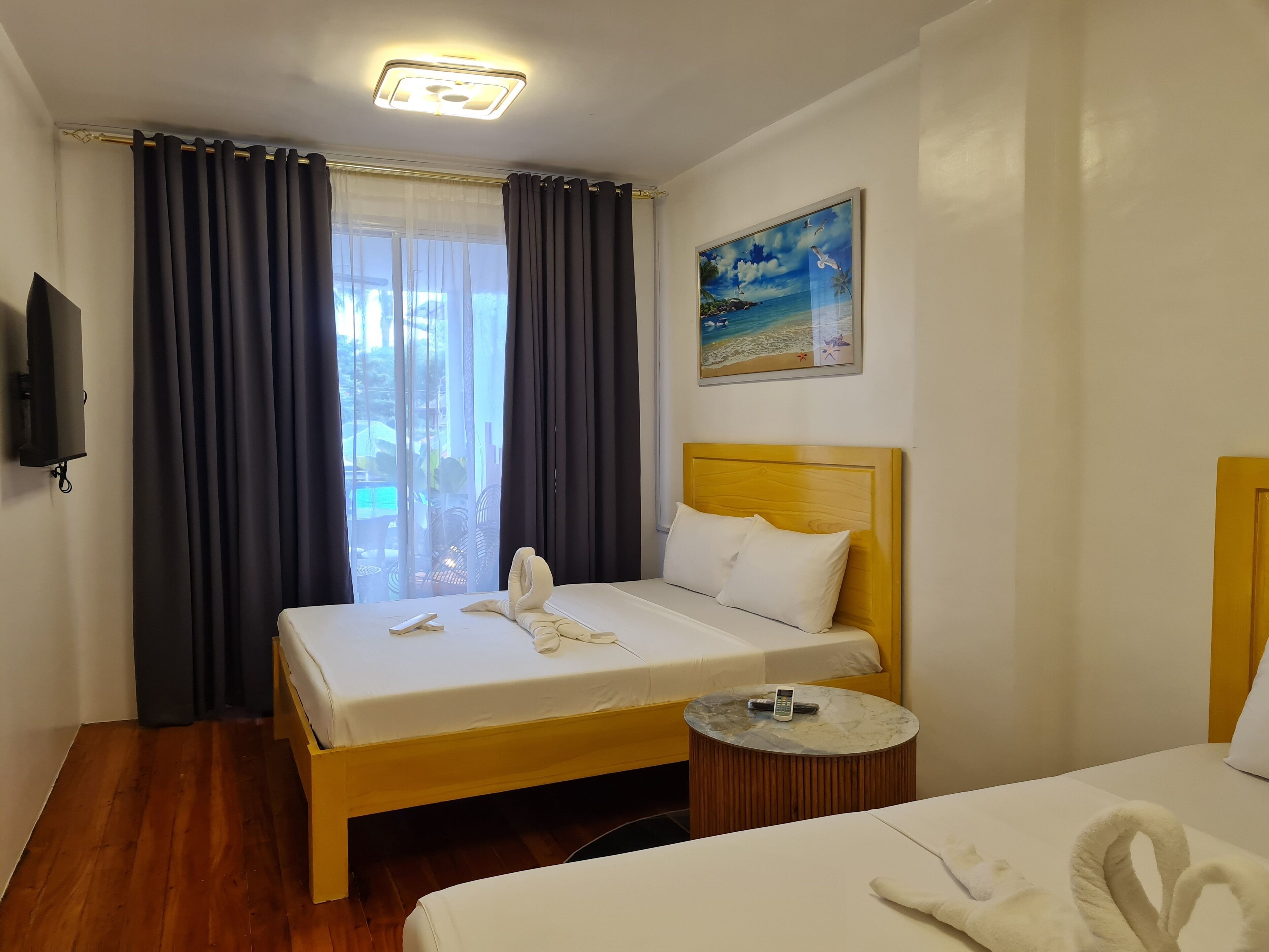 A hotel room with a bed, a round wooden side table, a painting of a beach scene, and a television mounted on the wall.