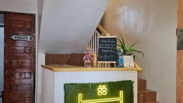 A hotel reception desk with a neon sign, a chalkboard with check-in/out times, and a wooden door labeled "TOILET".