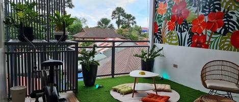 A balcony with a view of a house, a wall with a tropical floral mural, a small round table with a yellow cushion, and a wicker chair.