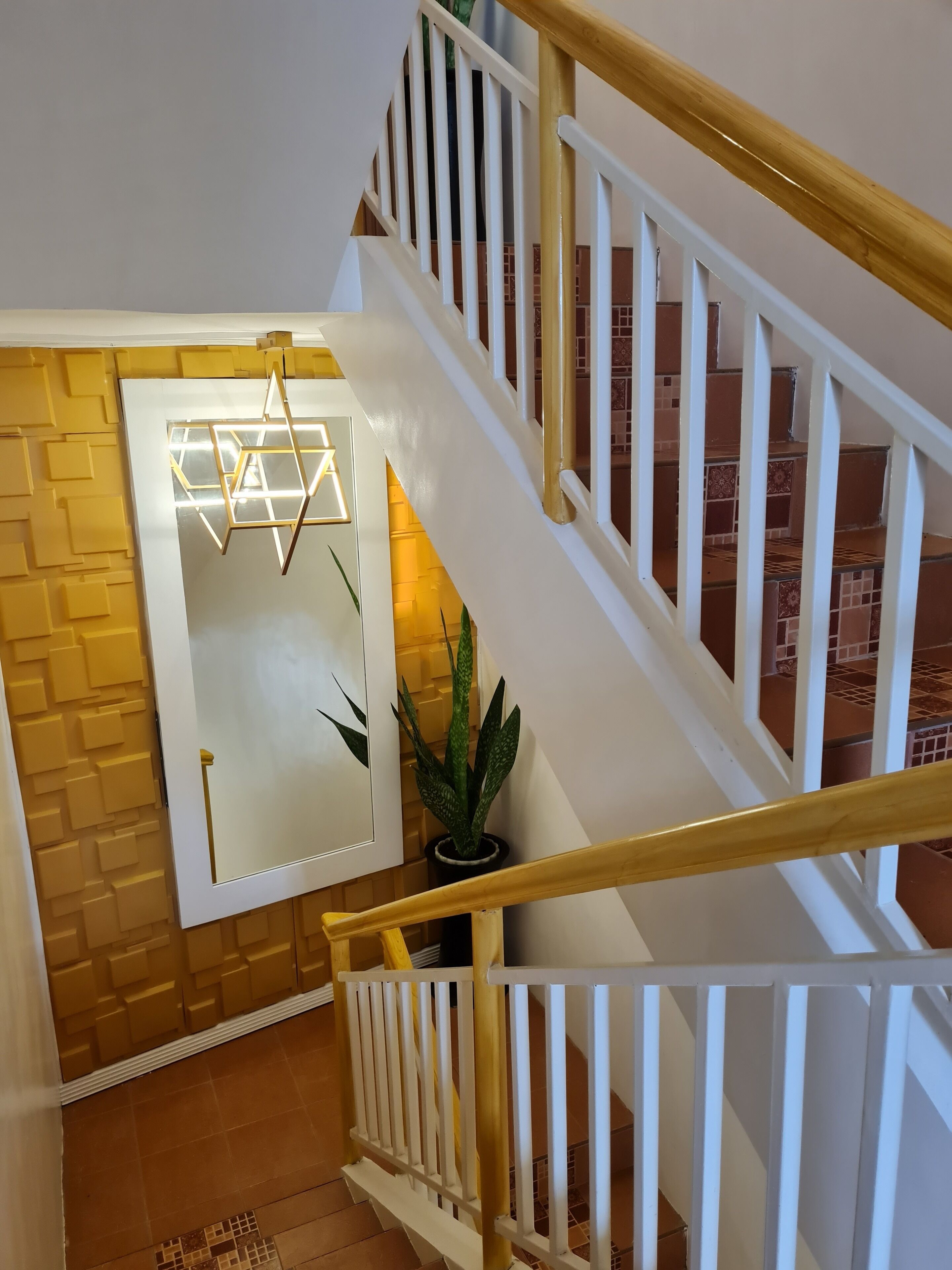 Een trap met witte leuningen en een houten handrail, die leidt naar een kamer met een wand met een geometrisch patroon en een potplant.