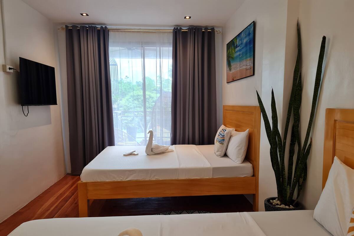A hotel room with a bed, a television, a window with curtains, a painting, and a potted plant.