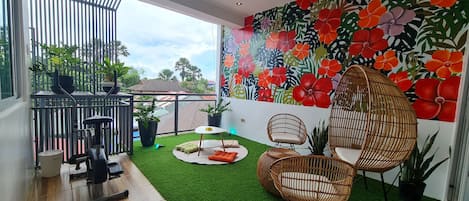 A balcony with a ceiling fan, exercise equipment, artificial grass, wicker chairs, and a vibrant floral mural.