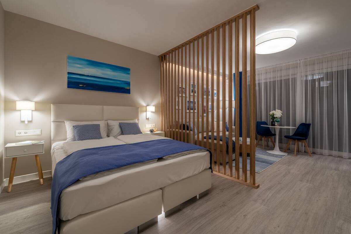 Ein modernes Hotelzimmer mit einem großen Bett, Nachttischlampen, einem Schreibtisch und Blick auf die Stadt durch transparente Vorhänge.