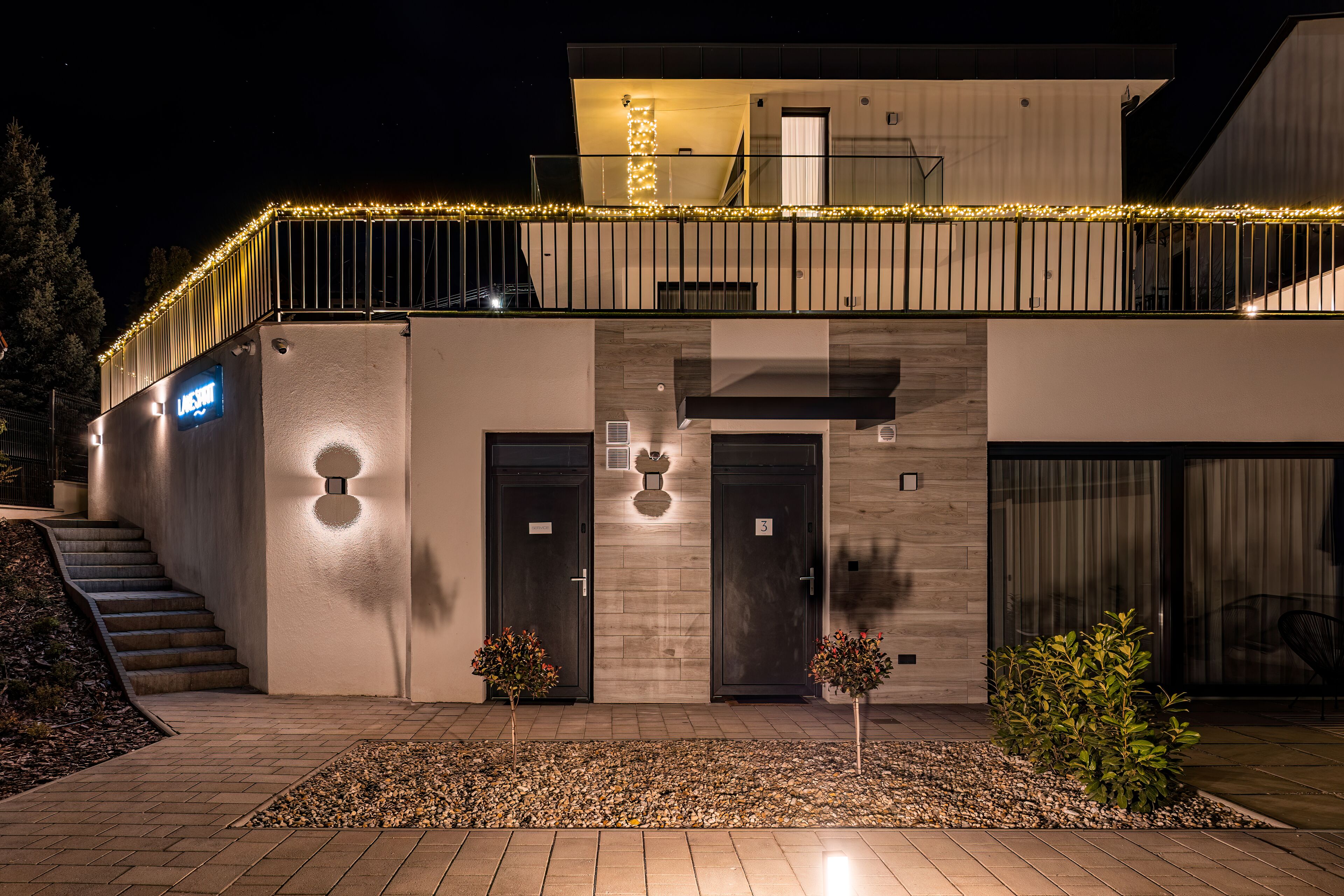 Un edificio moderno con un acceso de dos pisos, una escalera y un pequeño jardín con luces decorativas.