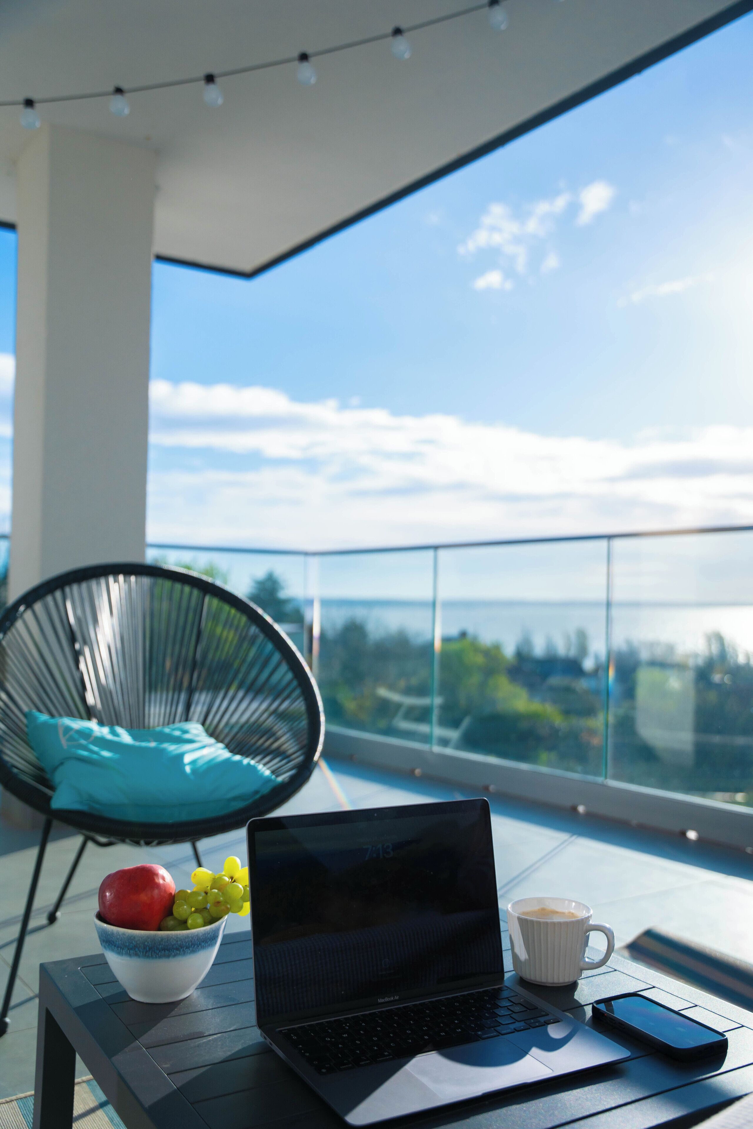 Una laptop sobre una mesa de balcón, con un frutero y una taza de café.