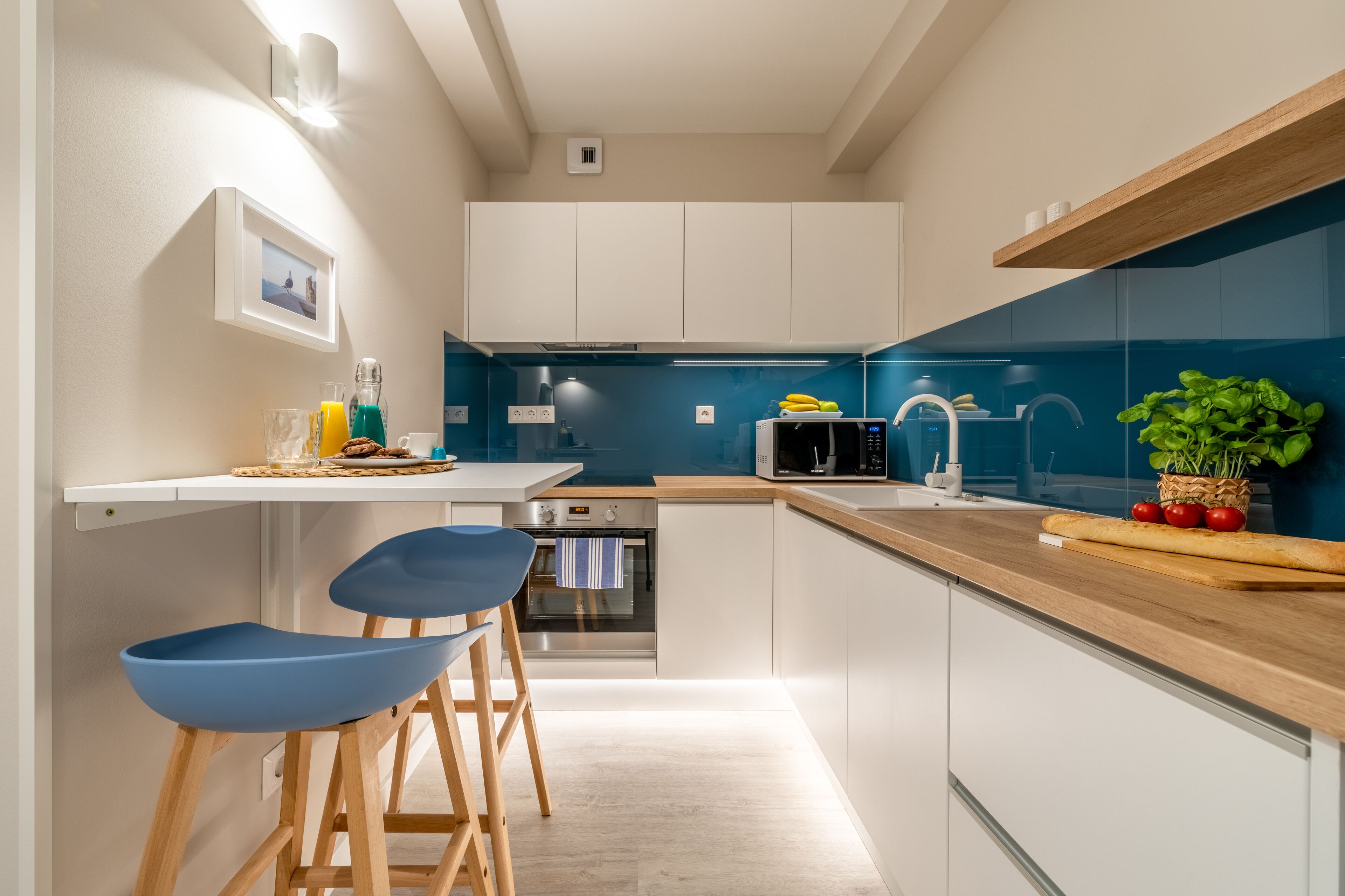 Una cocina moderna con armarios blancos, salpicadero azul y encimeras de madera. Hay dos taburetes de bar azules y un microondas.
