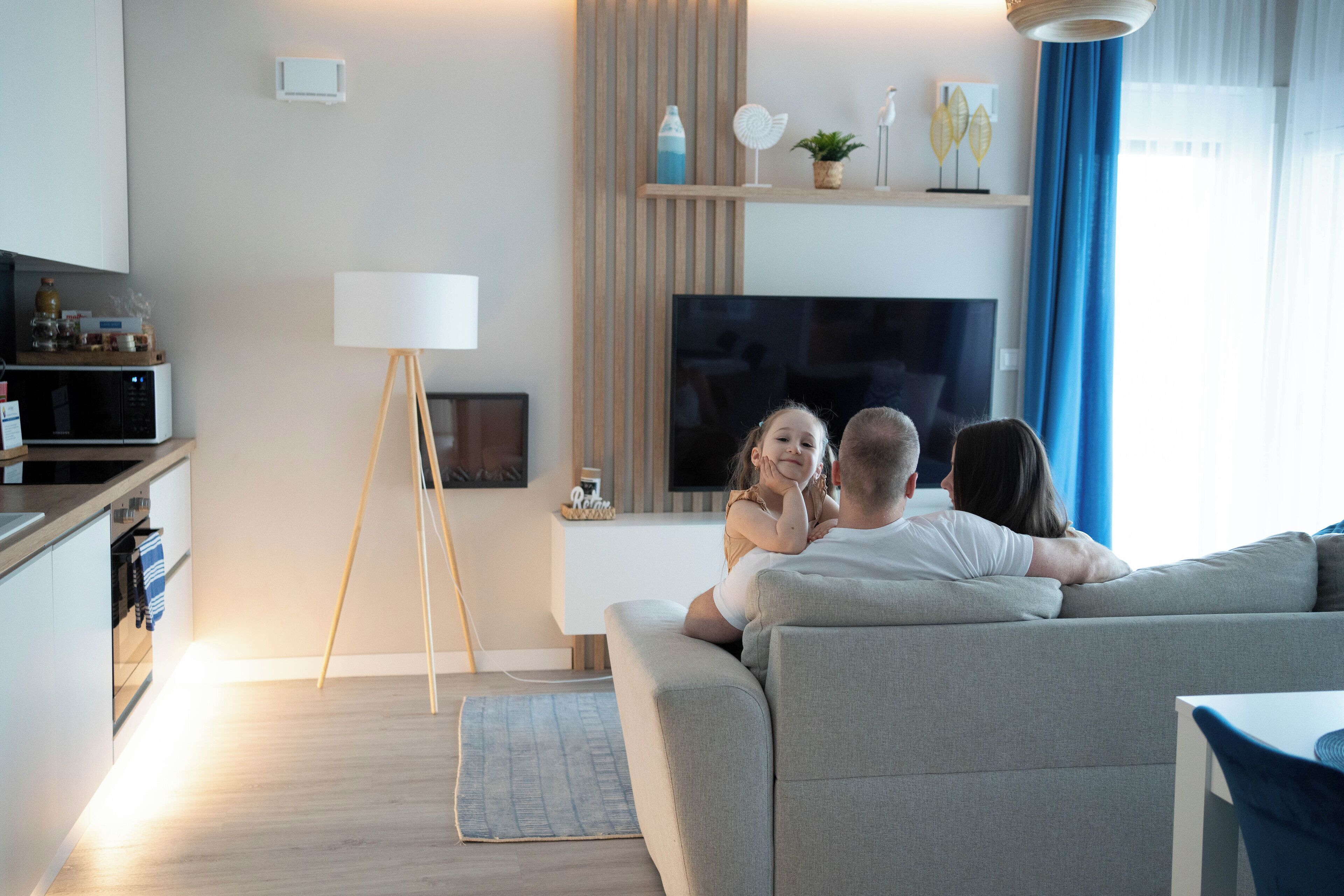A modern living room with a sofa, a flat-screen TV, a small table, and a kitchen area in the background.