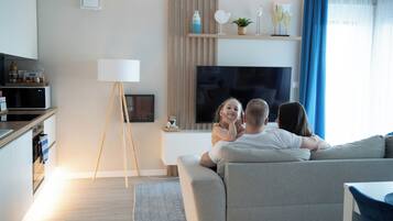 A modern living room with a sofa, a flat-screen TV, a small table, and a kitchen area in the background.