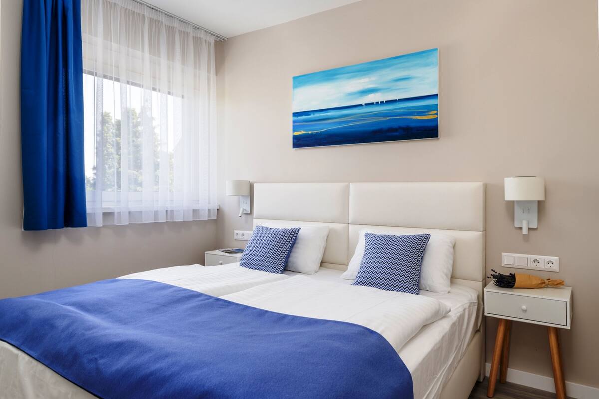 Ein Hotelzimmer mit einem großen Bett, einem Landschaftsbild und einem Fenster mit blauen Vorhängen.