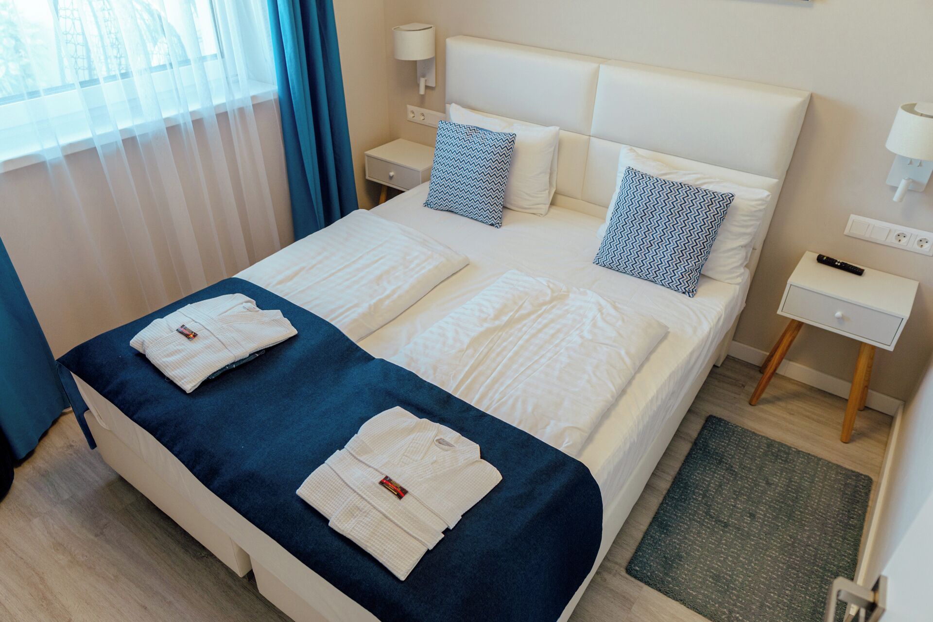 Une chambre d’hôtel avec un grand lit, deux tables de chevet, une petite table et une fenêtre avec des rideaux.