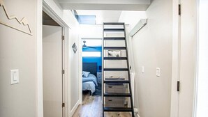 A narrow hallway with a black ladder leading to a bedroom with a bed and a blue wall. - Brooklyn Circle Loft — Newly Built 1BR + Loft, Hilltop, Walk Downtown (Leadville)