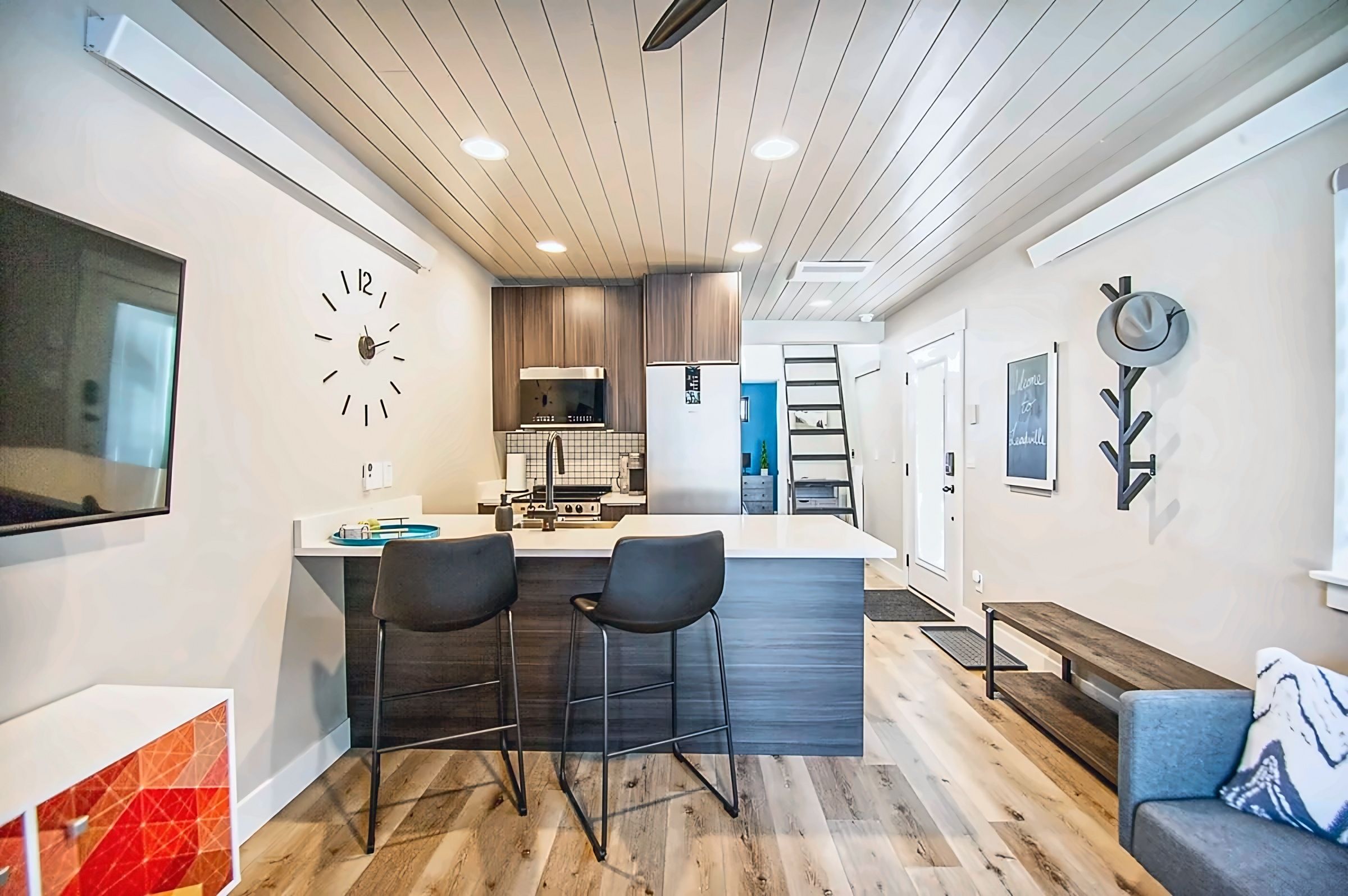 A modern kitchen with a central island, two bar stools, a clock, and a wall-mounted hat rack.
