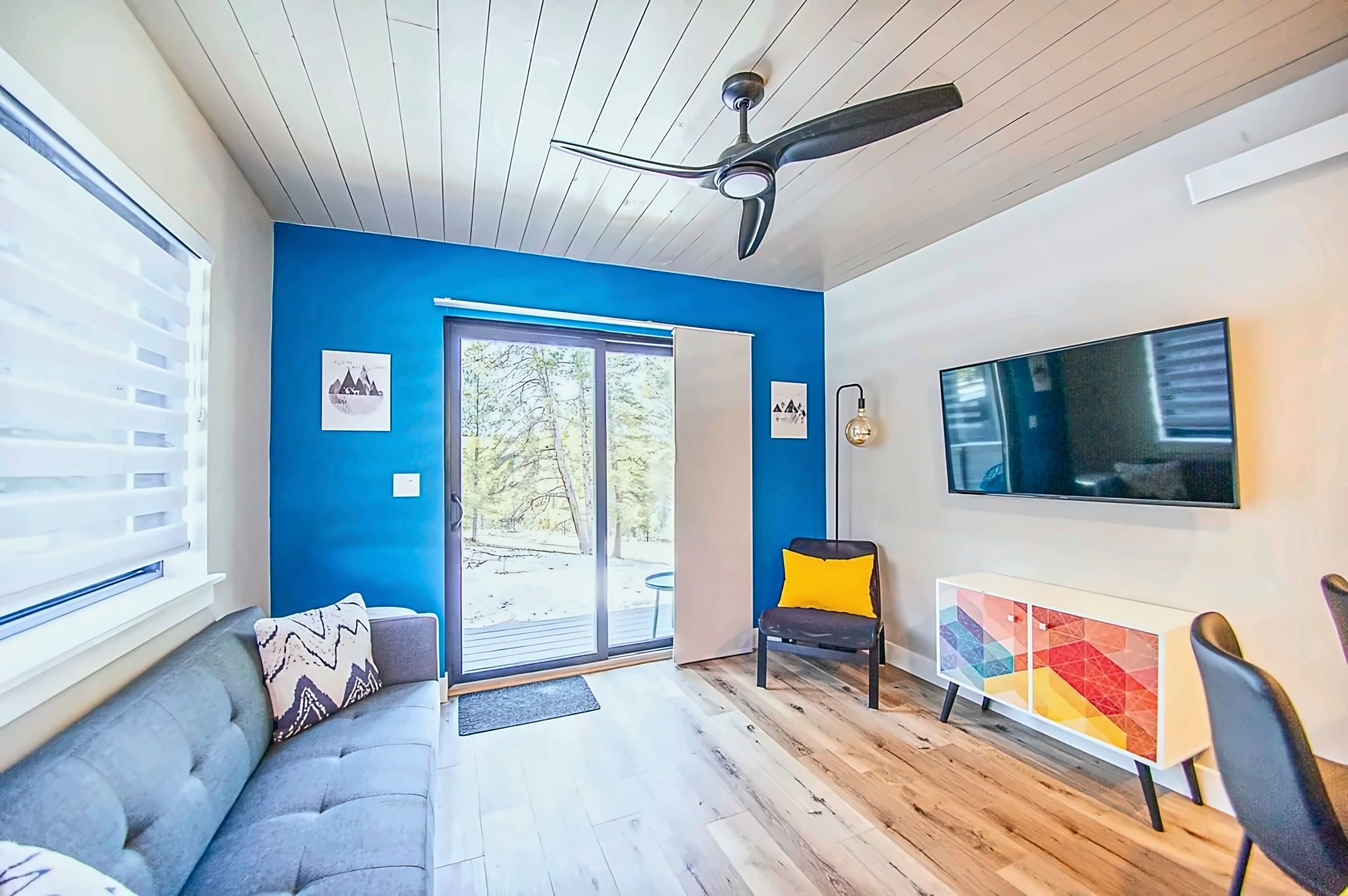 A modern living room with a blue accent wall, a ceiling fan, a sofa, a chair, a TV, and a glass door leading to an outdoor area.