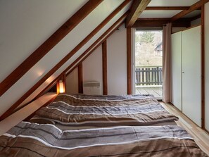 Une chambre avec un plafond en pente, un lit avec une literie à rayures, un radiateur et une vue sur une terrasse en bois par la fenêtre.